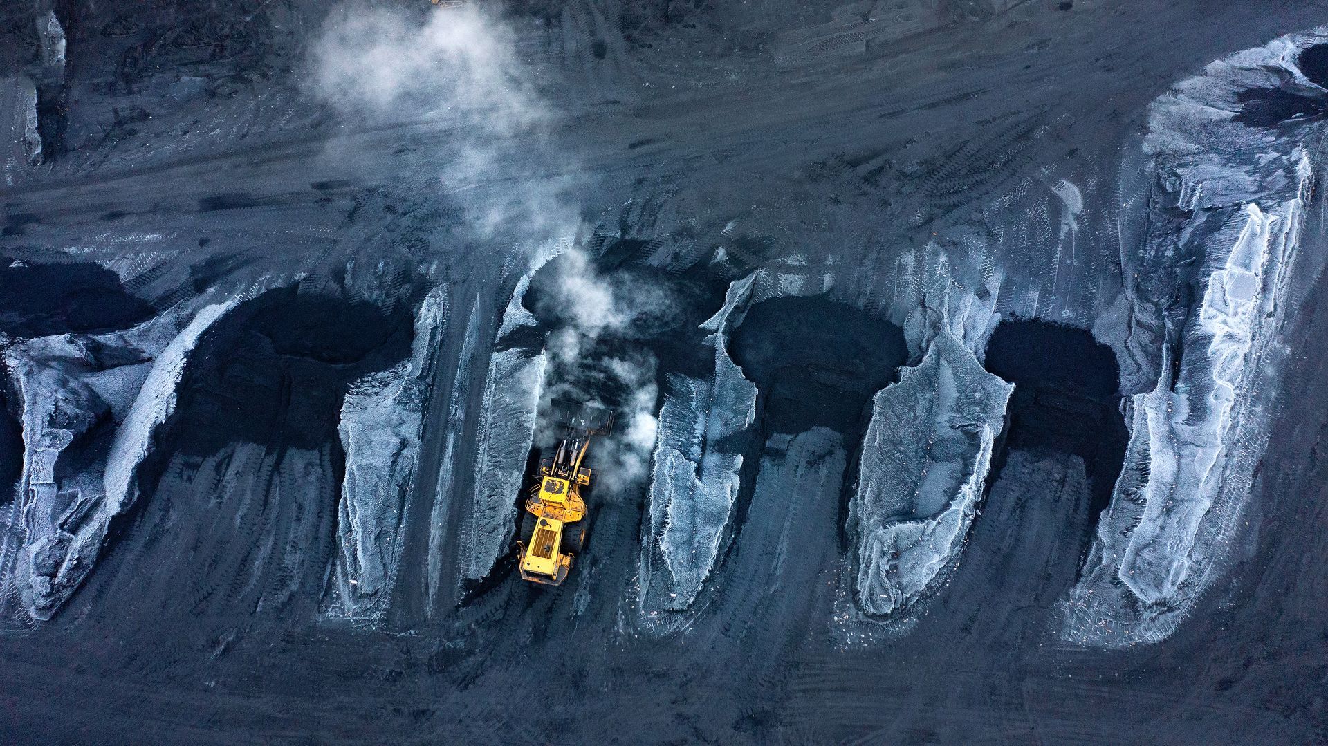 A front loader machine appears small and bright against the black piles of coal and coke in a manufacturing facility. Photographer used drone technology to create the overhead view of the manufacturing operations. 