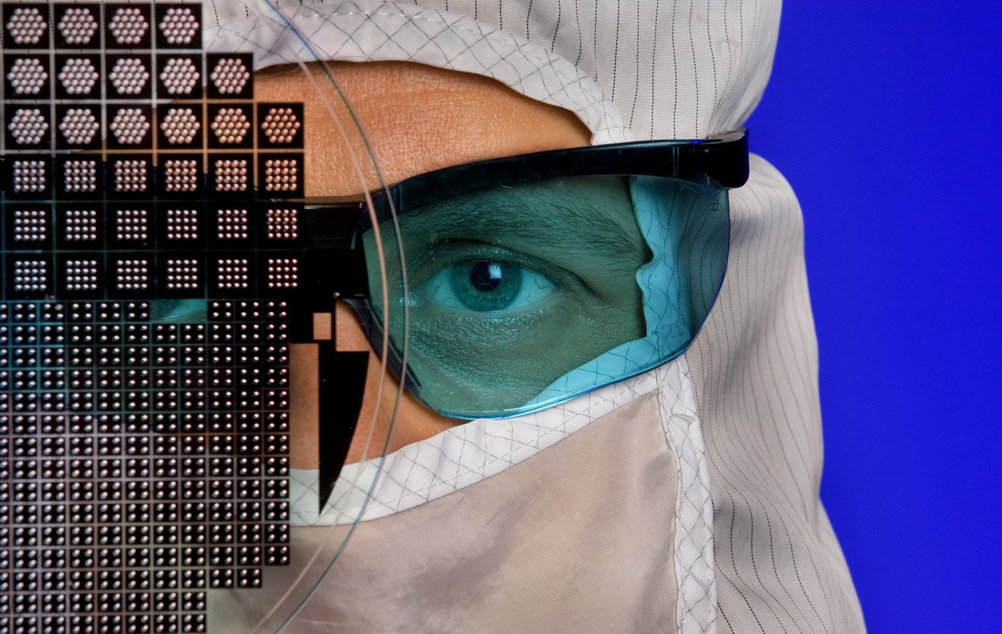 A worker in a clean room manufacturing facility holds a wafer chip up for the camera. The worker is looking through the wafer at the camera. It is a tight shot of the wafer and the worker's face. The worker is wearing a full-body clean suit. 