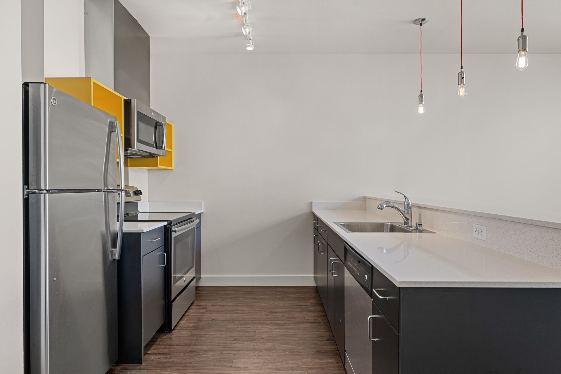 Modern kitchen interior with stainless steel appliances, light countertops, and dark cabinetry against a yellow backsplash. Features include a refrigerator, dishwasher, and microwave. There are exposed filament pendant lights hanging above the sink area. The room has wood flooring and neutral wall colors at Anthem on 12th in Seattle, WA.