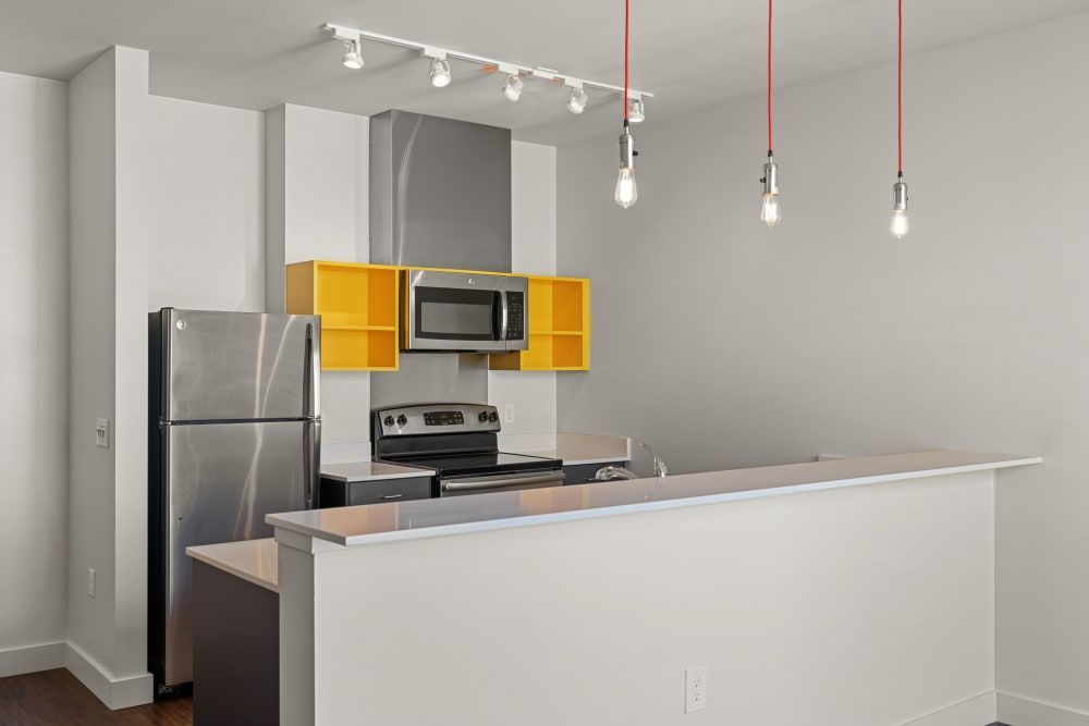 Modern kitchen interior with gray and white walls, featuring stainless steel appliances including a refrigerator and an electric range with an overhead microwave. Bright yellow upper cabinets add a pop of color above the stove and microwave. A white countertop with a sink is visible, and red pendant lights hang from the ceiling, providing accent lighting. The space is clean and appears new or recently renovated at Anthem on 12th in Seattle, WA.