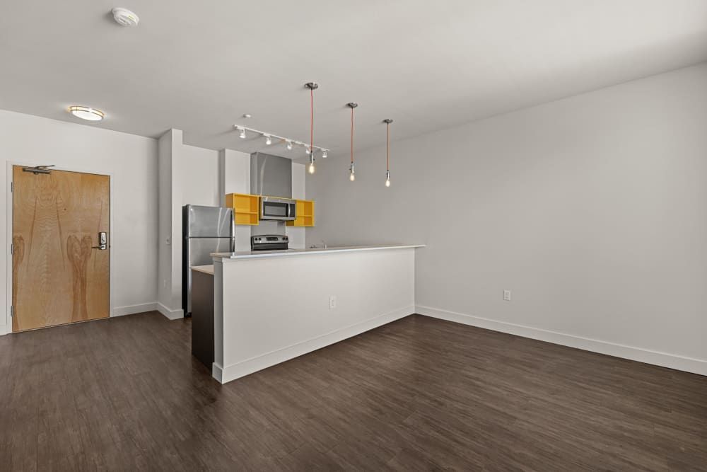 Modern kitchen with white and yellow cabinets, stainless steel appliances, and a breakfast bar, opening into a spacious, empty living area with dark wood flooring, white walls, and recessed lighting at Anthem on 12th in Seattle, WA.