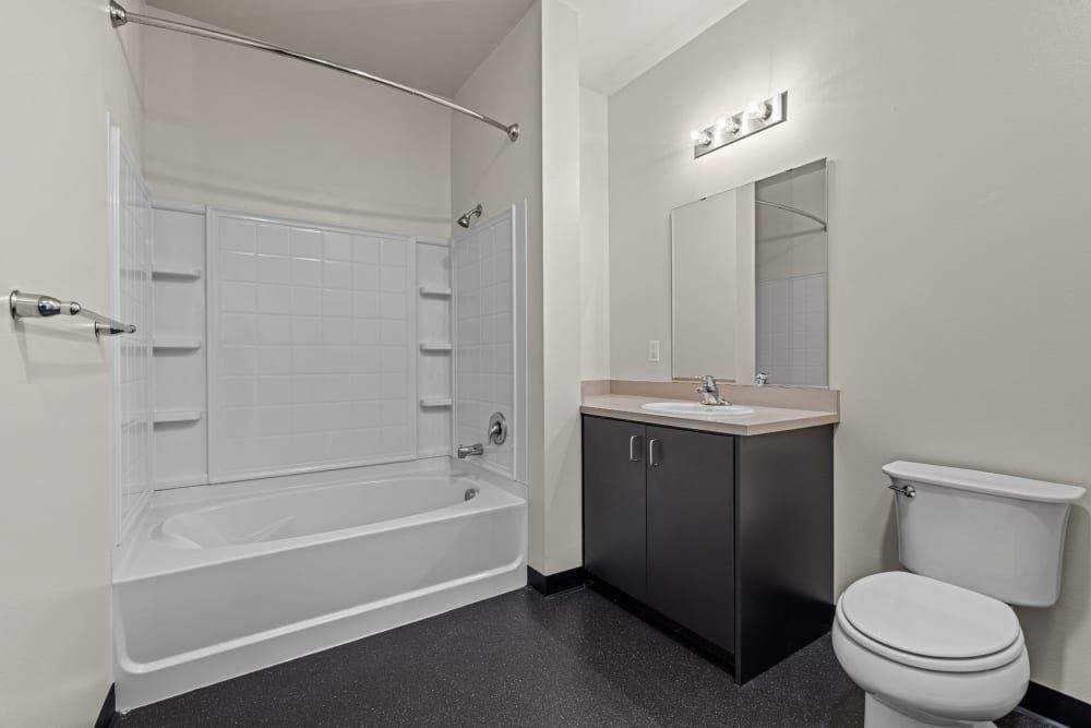 Modern bathroom interior with a white bathtub and shower combination on the left with a clear glass sliding door and mounted towel bars. To the right, there's a dark wood vanity with an undermount sink and a mirror above, flanked by a white toilet. The flooring is speckled, and the room is painted in a neutral tone at Anthem on 12th in Seattle, WA.