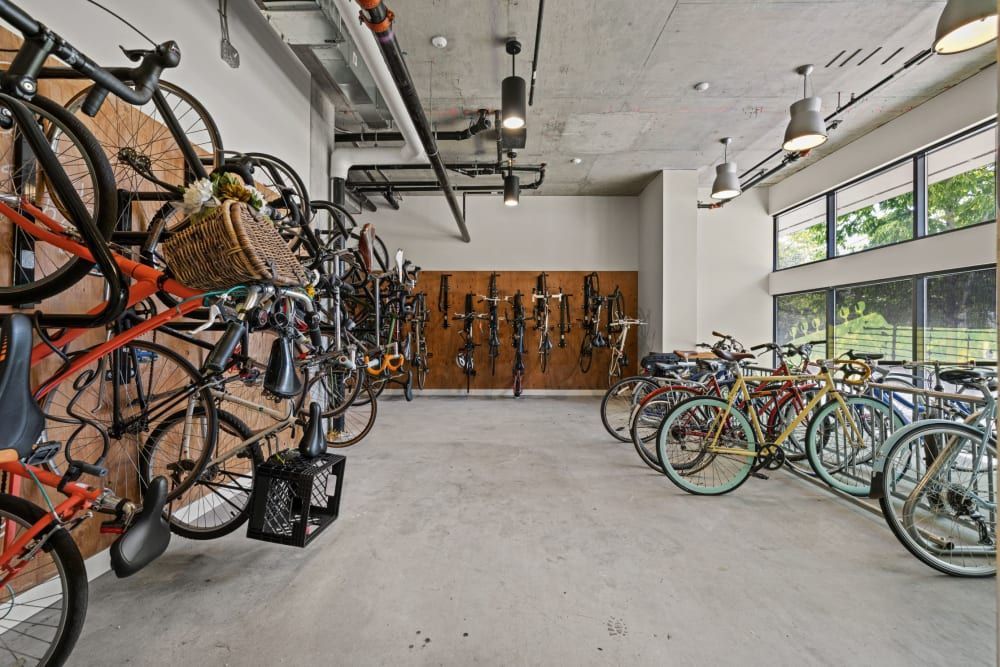 Interior of a modern bicycle parking room with multiple bikes stored vertically on wall-mounted racks to the left and horizontally on floor stands to the right. The room is well-lit with pendant lights and natural light from large windows, and has a polished concrete floor and exposed ceiling infrastructure at Anthem on 12th in Seattle, WA.
