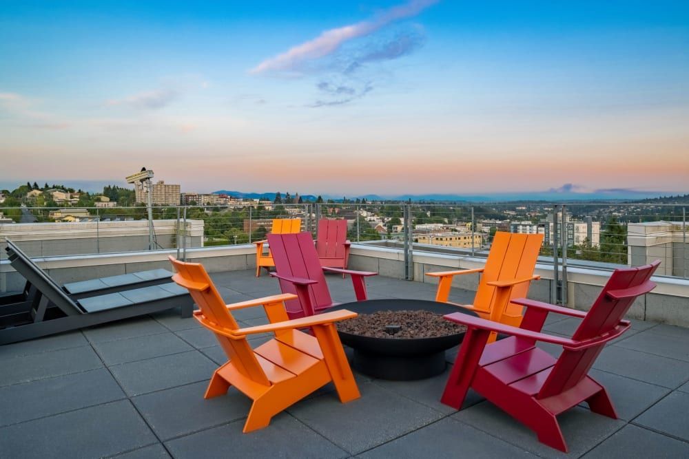 A rooftop terrace at dusk featuring a fire pit surrounded by bright orange and red Adirondack chairs, with a scenic view of a hilly landscape and buildings under a wide sky showing warm hues and a few wispy clouds at Anthem on 12th in Seattle, WA.