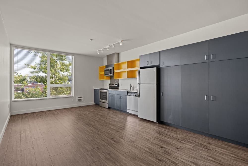 Modern kitchen in an apartment with gray cabinets, stainless steel appliances, and yellow upper cabinet accents. The room has laminate flooring, a large window with a view of greenery, and track lighting on the ceiling at Anthem on 12th in Seattle, WA.