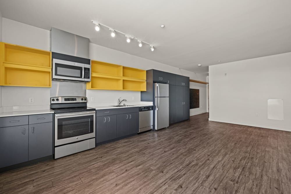 Modern kitchen interior with dark gray lower cabinets and yellow upper cabinets, stainless steel appliances, including a refrigerator, dishwasher, stove, and microwave, with track lighting on the ceiling and wood laminate flooring extending into an open living space with white walls at Anthem on 12th in Seattle, WA.