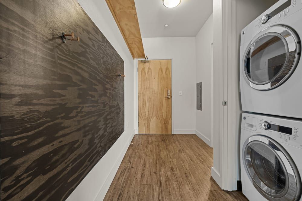 Interior view of a modern laundry room with stacked washer and dryer to the right. The room features wood-look flooring, white walls, and a sliding barn door on the left side with a unique wooden texture. Recessed lighting is visible on the ceiling and a wooden door can be seen in the background at Anthem on 12th in Seattle, WA.