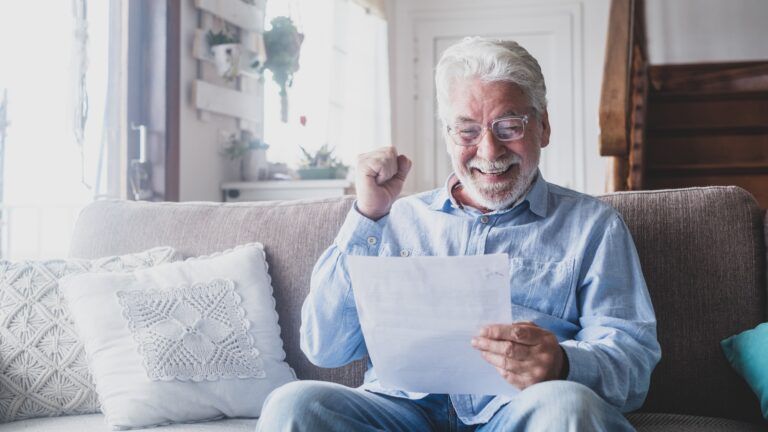 An elderly man is sitting on a couch holding a piece of paper.