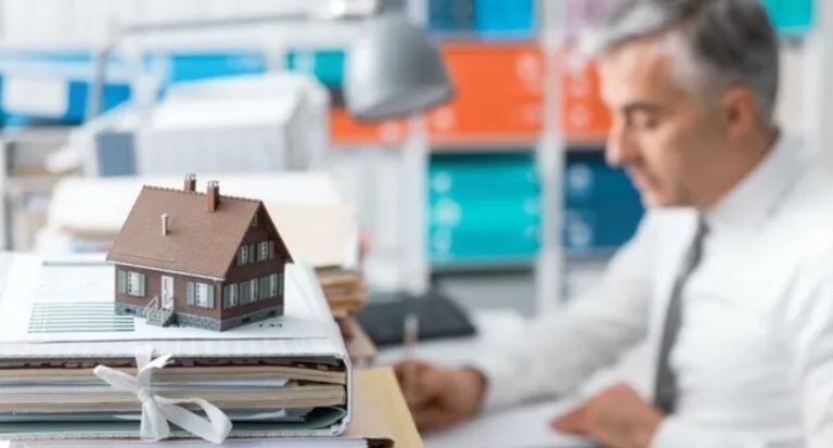 A man is sitting at a desk with a model house on top of a pile of binders.