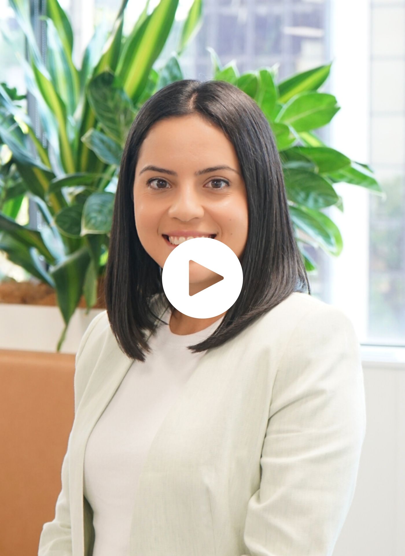 A woman in a white jacket is smiling in front of a plant.