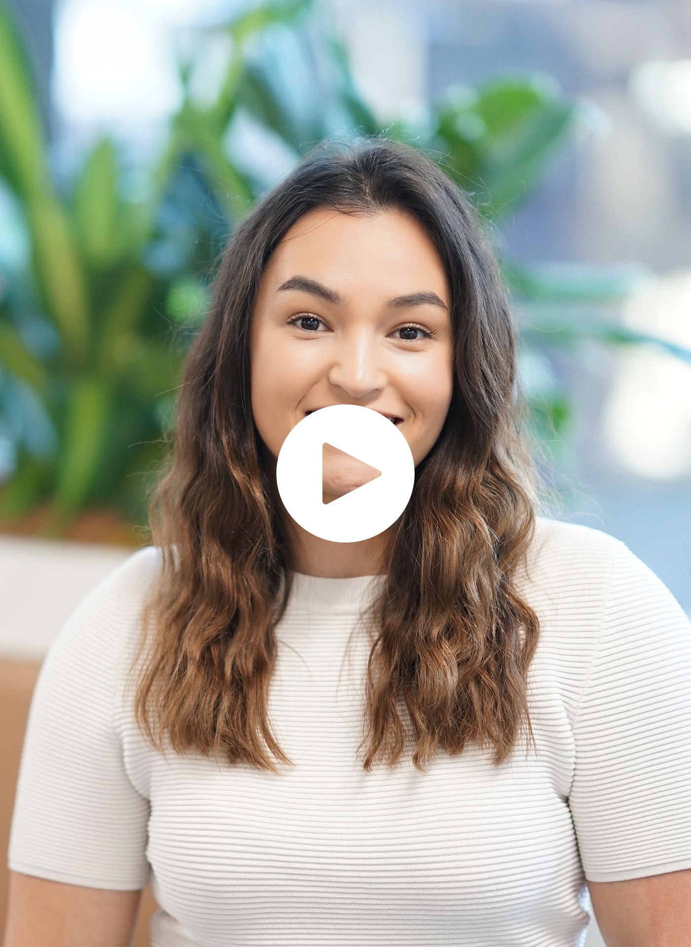 A woman in a white shirt is smiling with a play button in front of her face.
