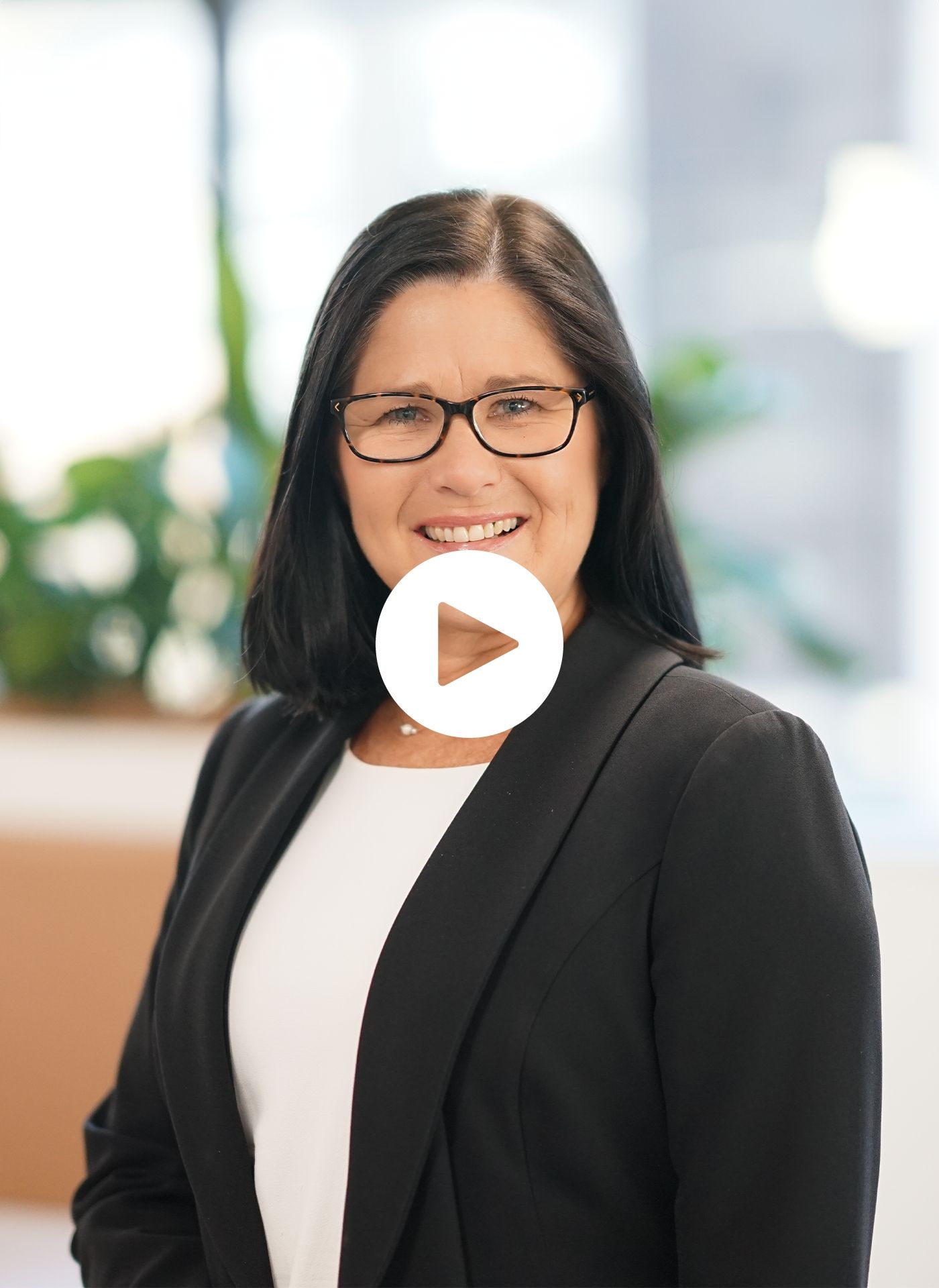 A woman wearing glasses and a black jacket is smiling in front of a play button.