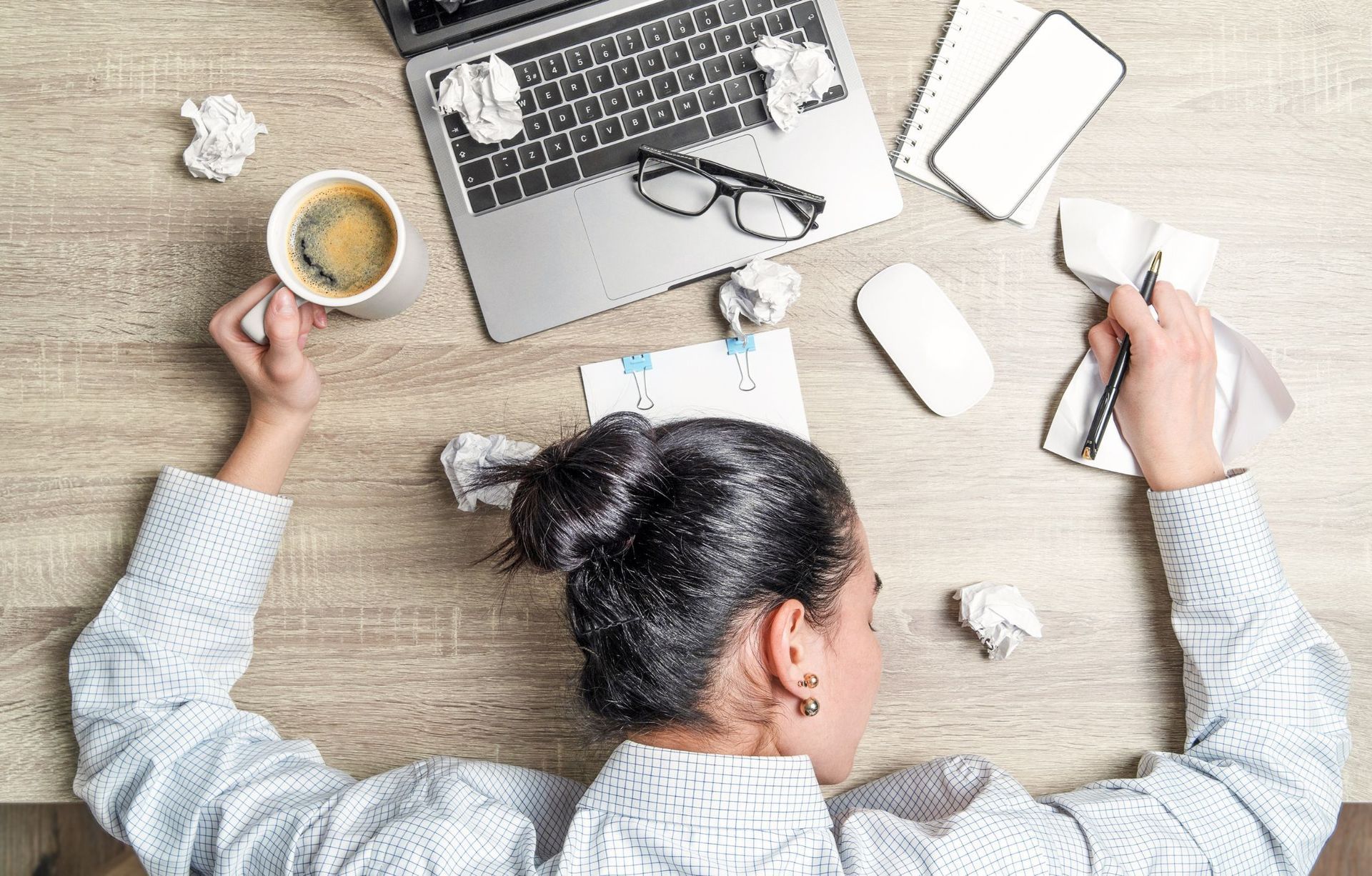 A woman is laying on a desk with a cup of coffee and a laptop.