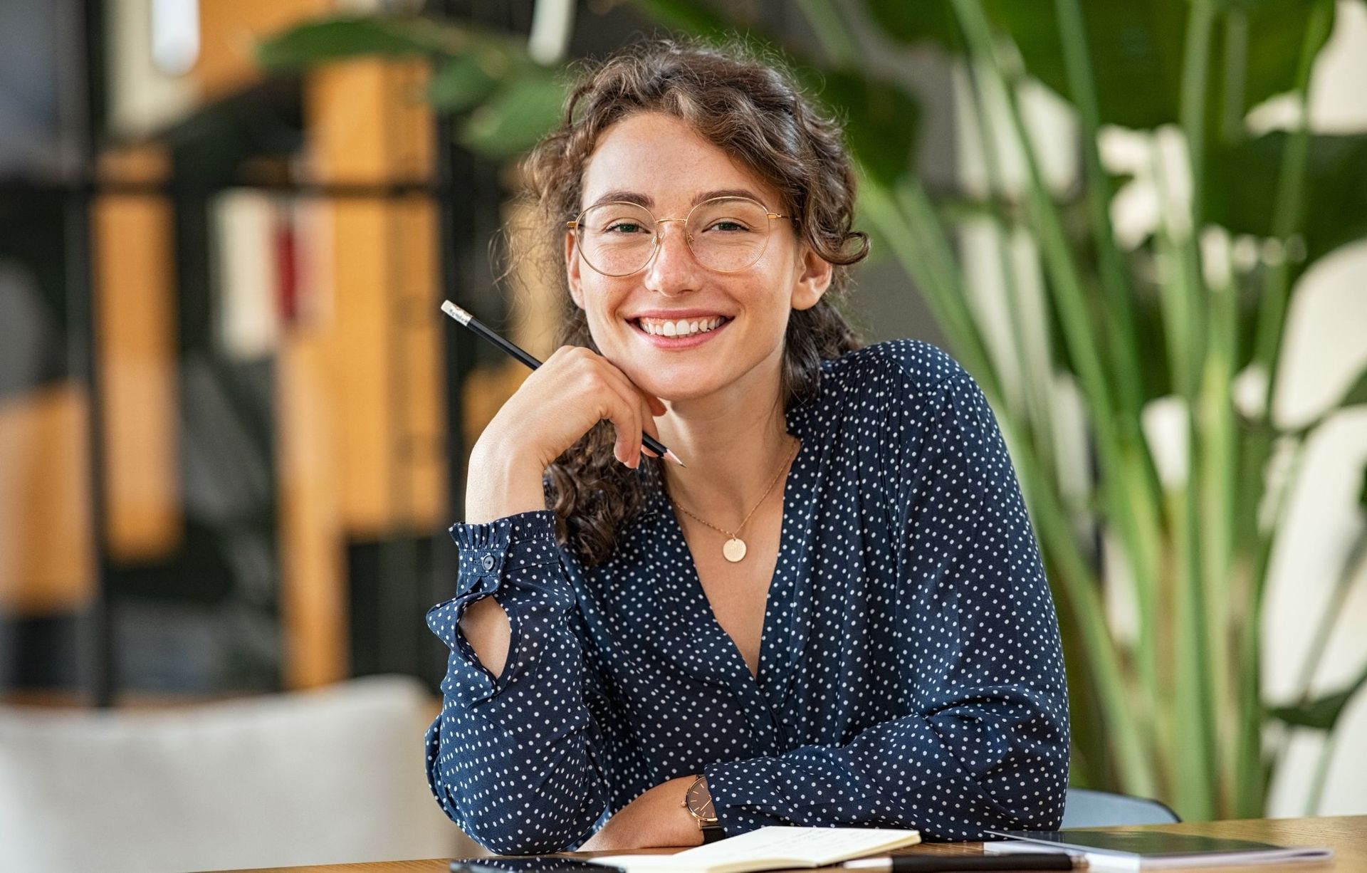 A woman is smiling while sitting at a table with a pen in her hand.