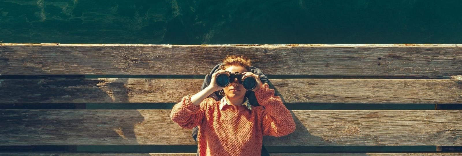 A woman is looking through binoculars on a wooden dock.