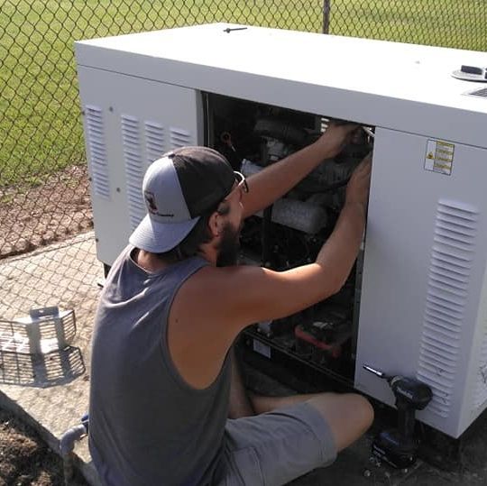 Person kneels beside an open white generator, adjusting its internal components outdoors.