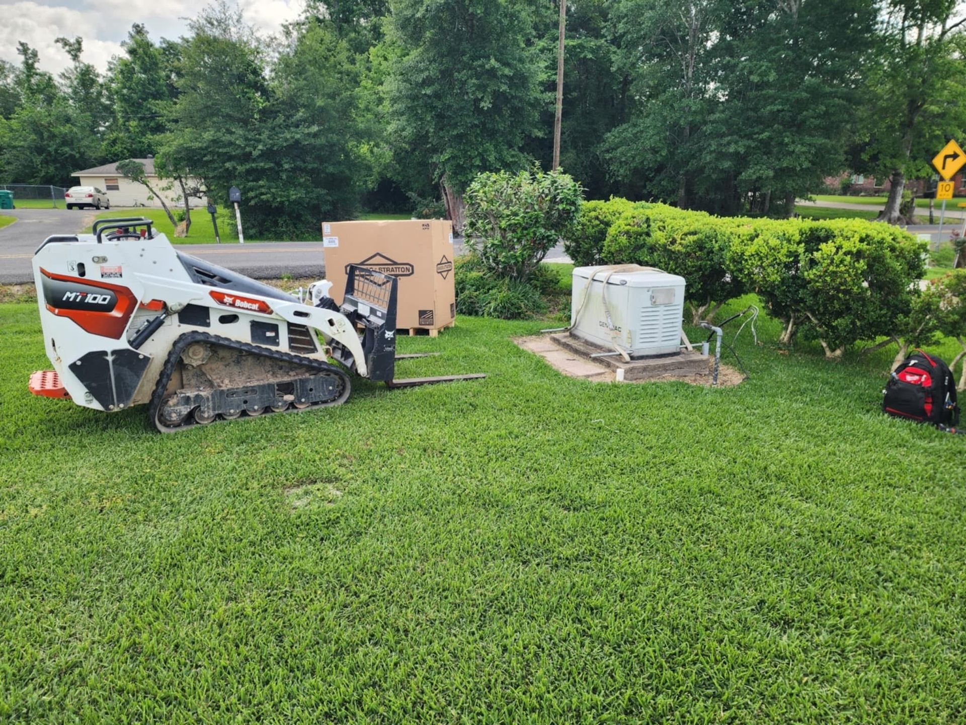 Compact skid steer and utility boxes on a grassy roadside near trees and a warning sign