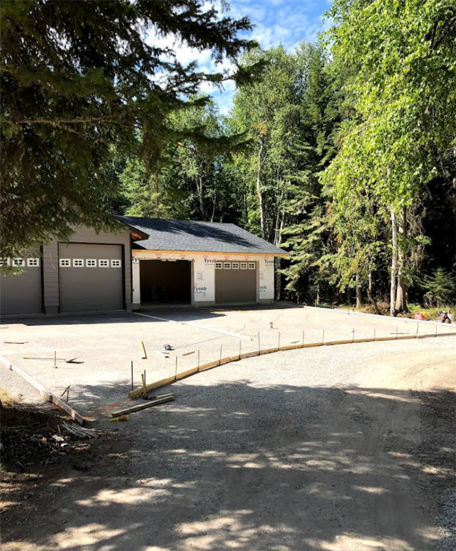 A garage with two garage doors is surrounded by trees and a dirt road.