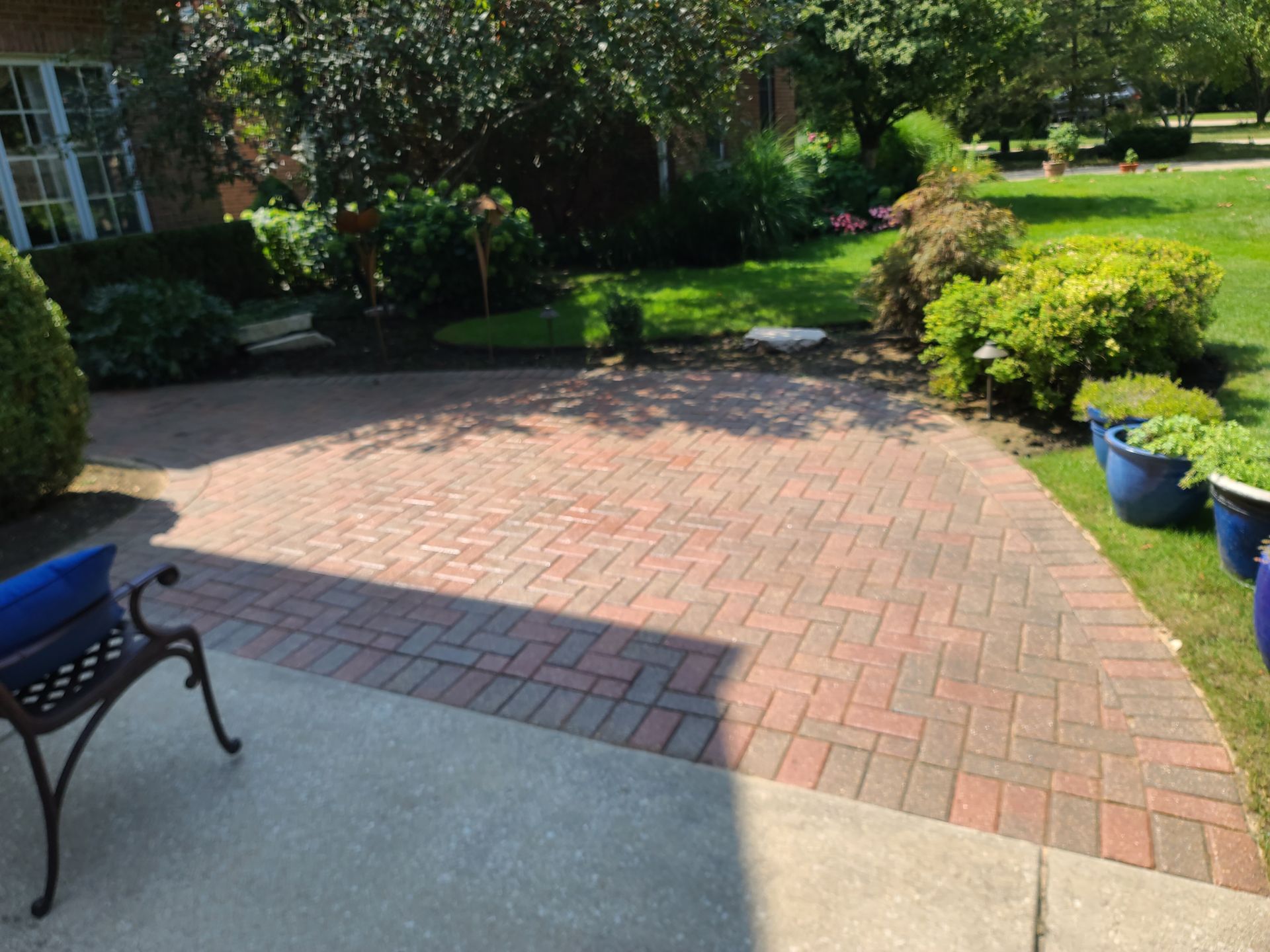 Brick patio in a yard with grass, bushes, and potted plants. Shadow from trees.
