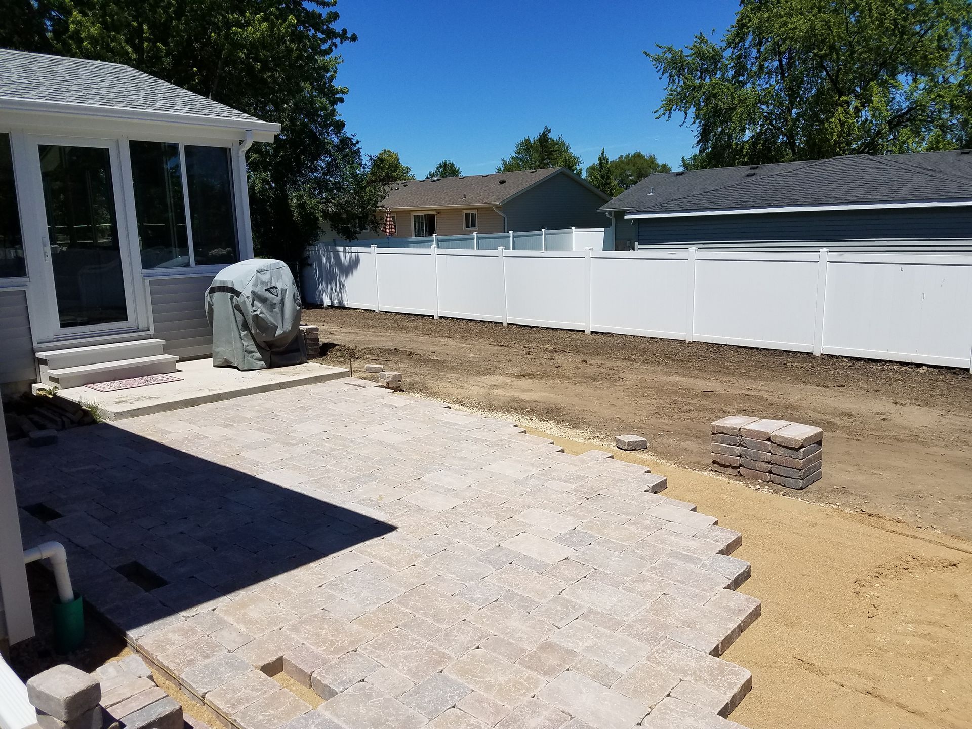 Backyard patio under construction with tan pavers, dirt, white fence, and house.