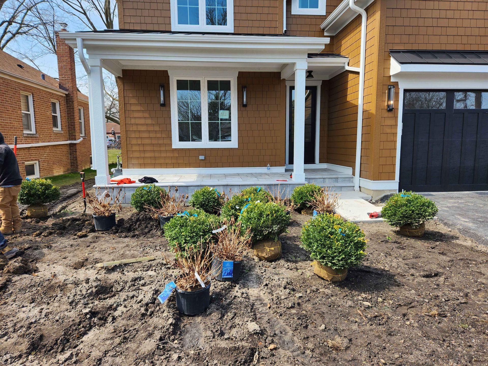 Front yard landscaping of a brown house with shrubs in pots on bare soil.