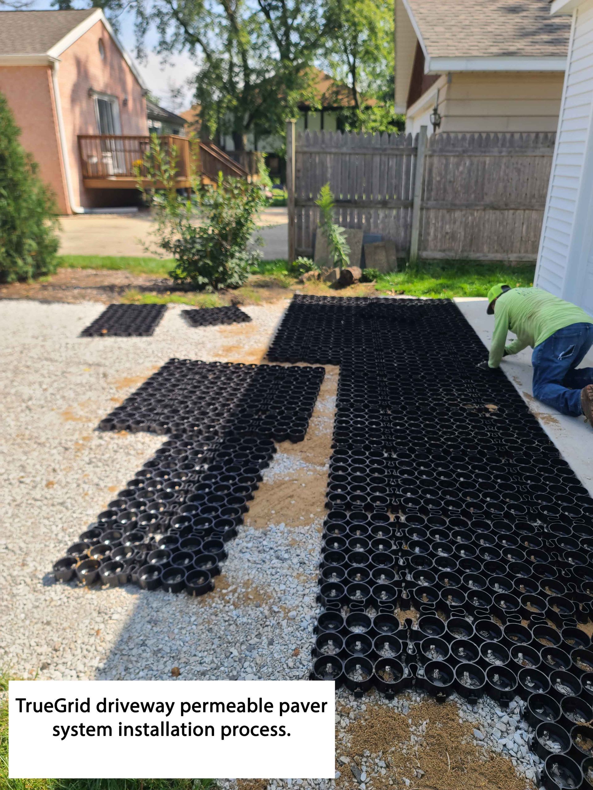 Person installing black permeable pavers on a gravel driveway in a yard.