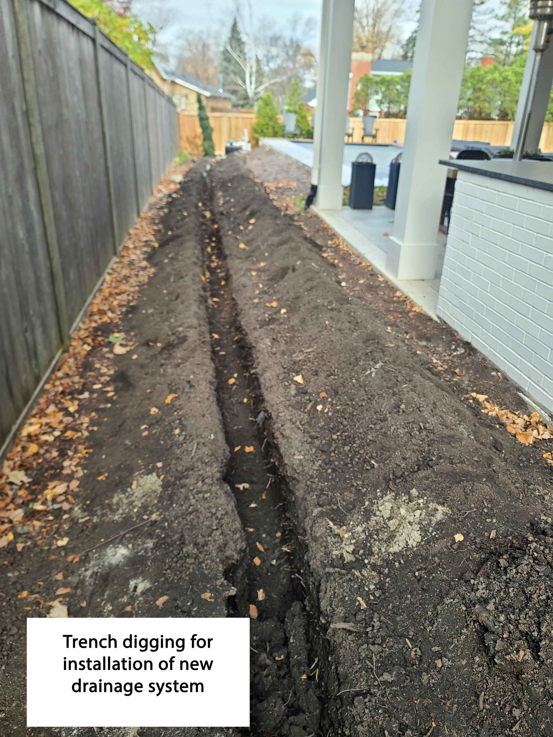 A long trench dug along a backyard fence for the installation of a new drainage system.