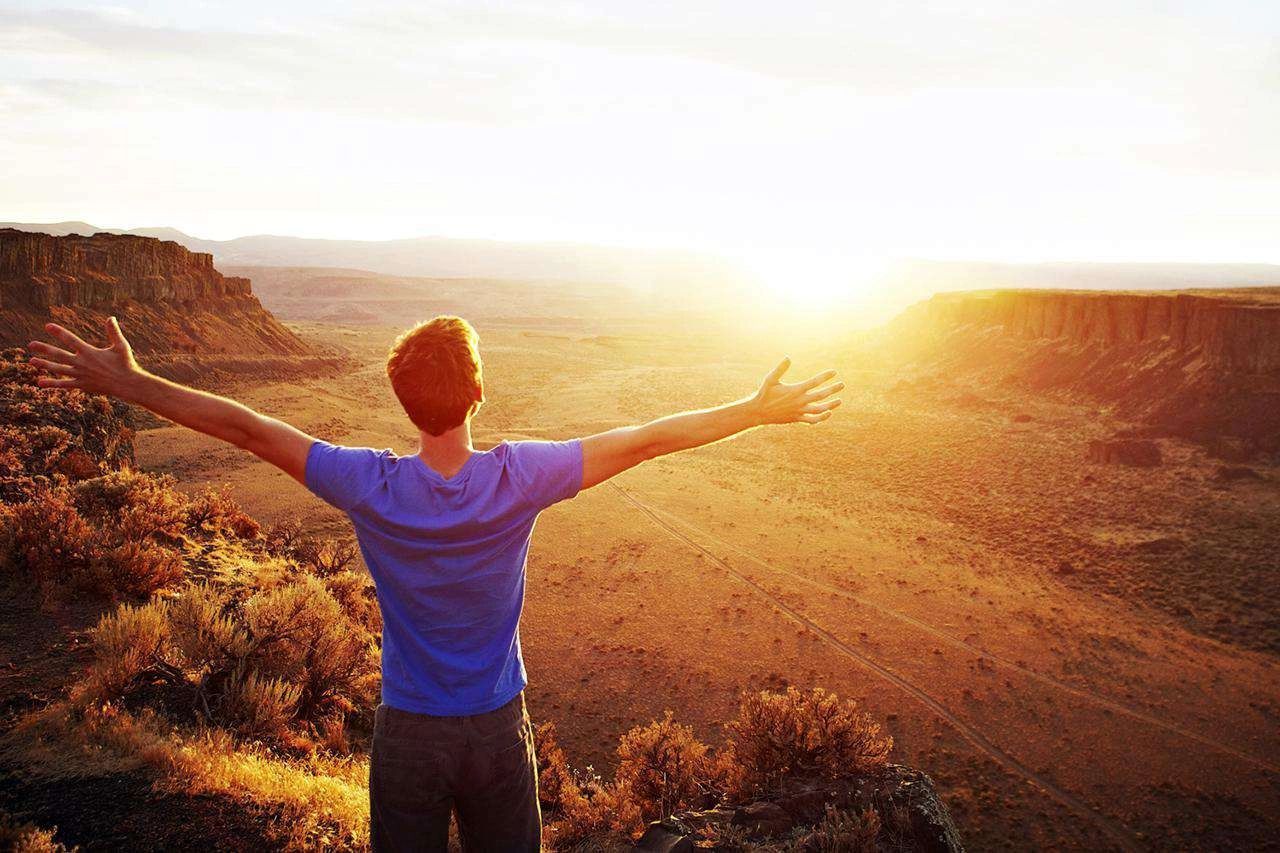 Person with arms outstretched, facing sunset over canyon.