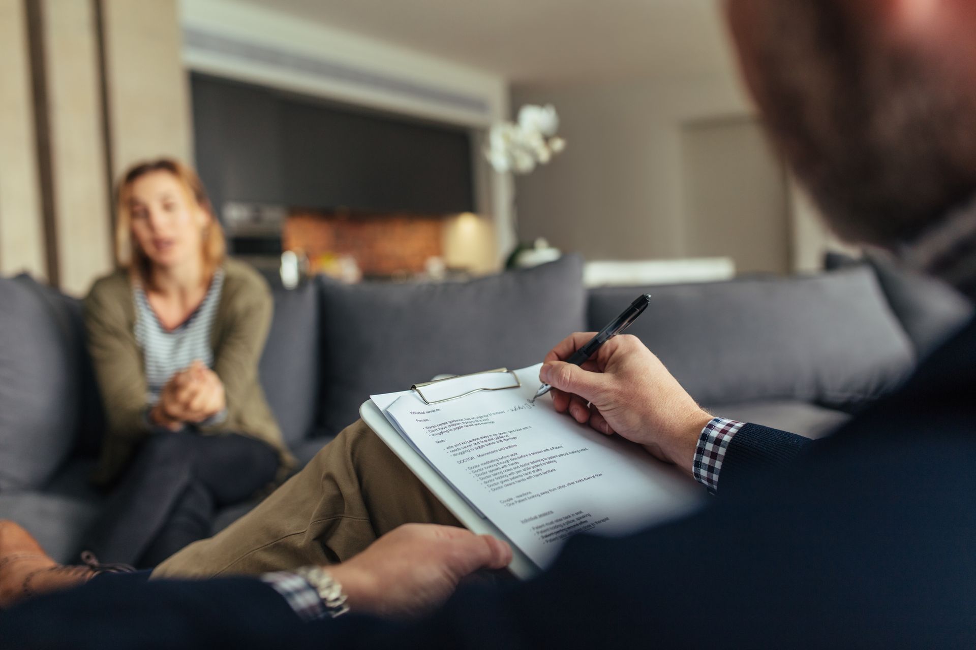 Therapist writing notes during a session with a patient seated on a couch. Living room setting.