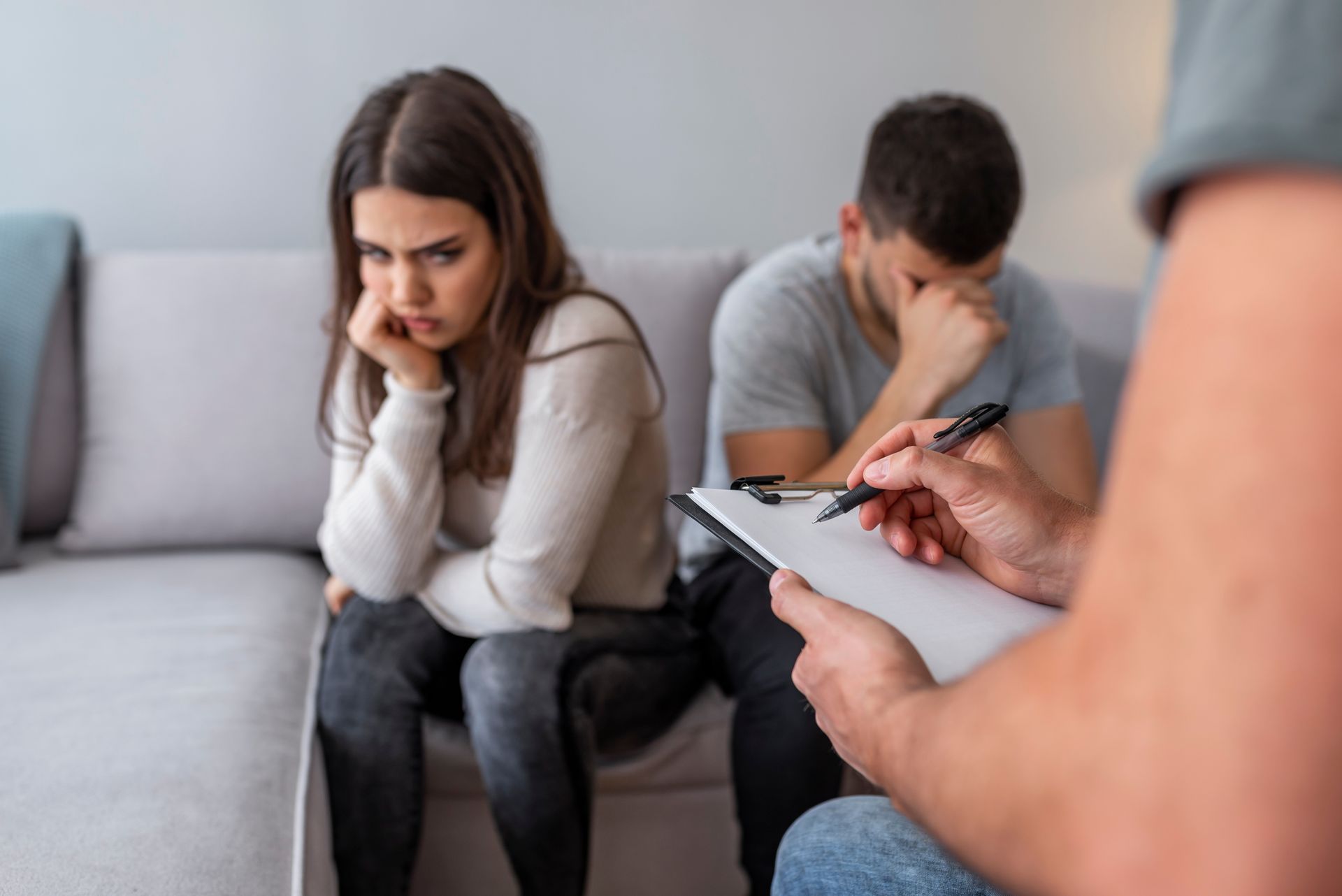 Couple in counseling session, facing away, with worried expressions, therapist taking notes.