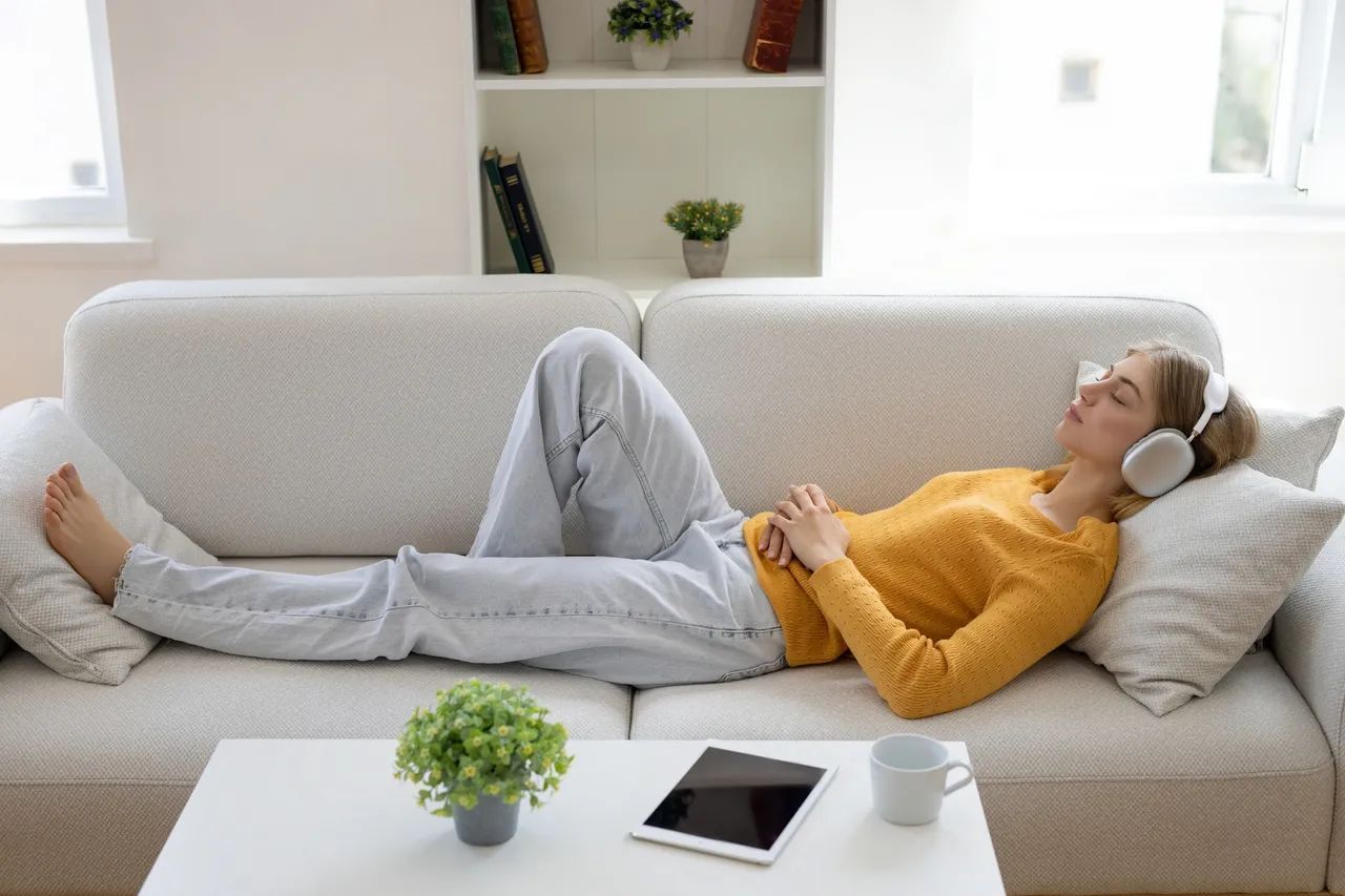 Woman wearing headphones resting on a couch, eyes closed. Table with tablet and plant in front.