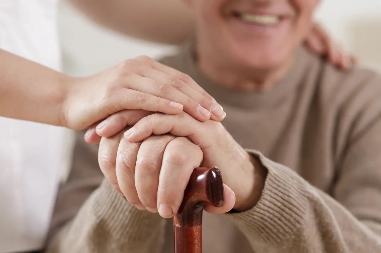 Person's hands held by another's, resting on a cane. One person smiles.
