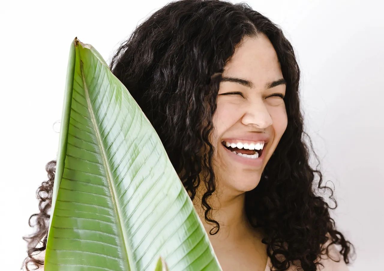 Woman with curly hair, smiling widely, holding a large green leaf, against a white background.