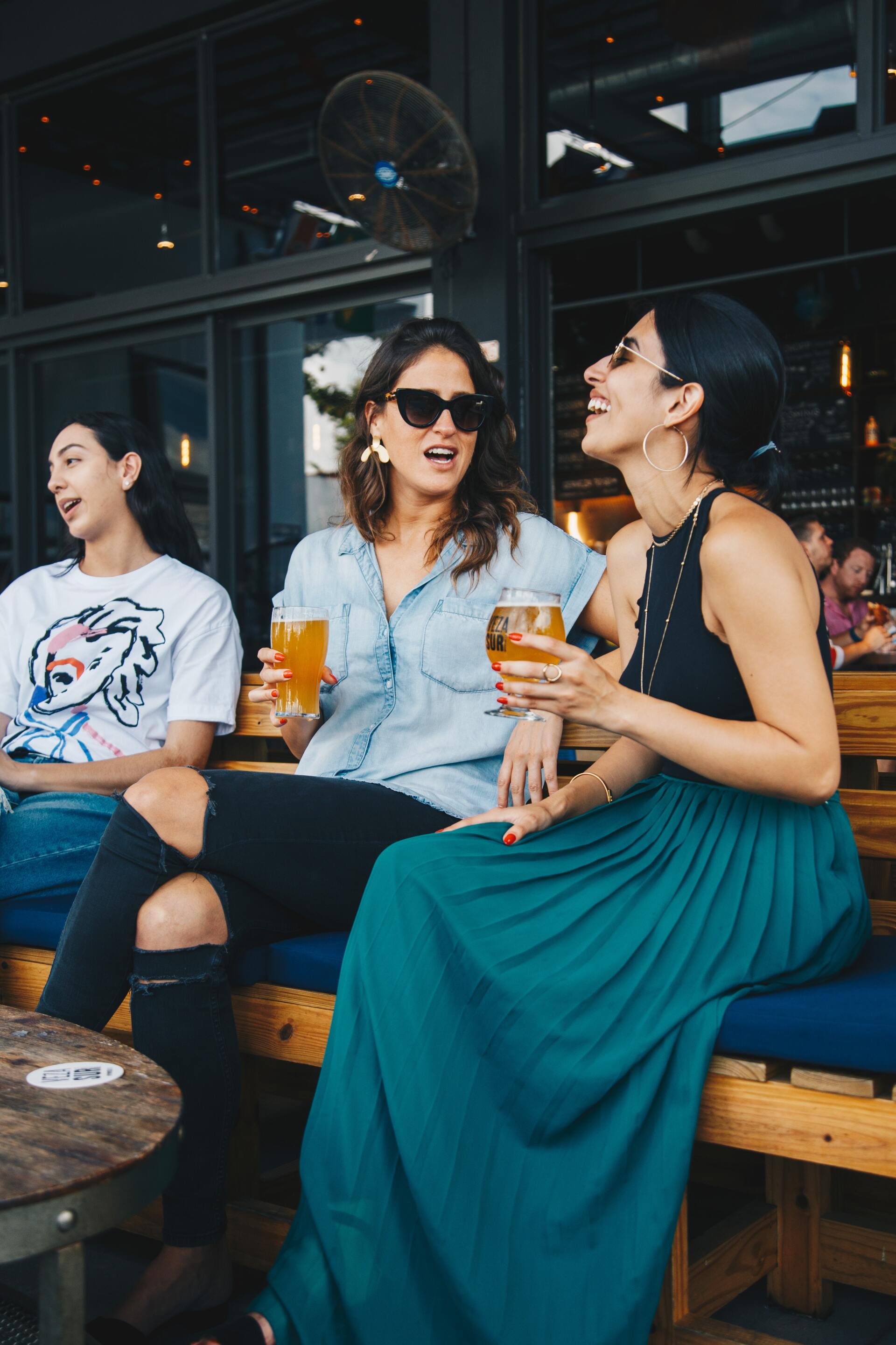 Tres mujeres están sentadas en un banco bebiendo cerveza y riendo.