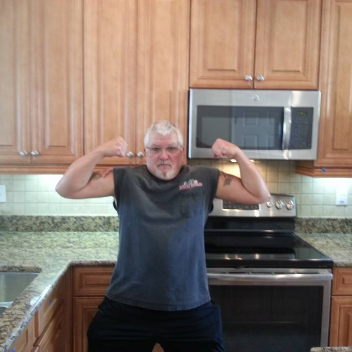 Man flexing biceps in a kitchen with wooden cabinets and stainless steel appliances.