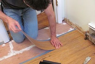 Person installing flooring: kneeling, placing a flexible plank over existing floor in a room.