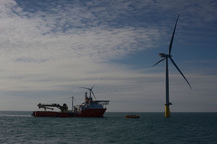 Offshore wind turbine with a large support vessel and small boat nearby on the water under a partly cloudy sky.