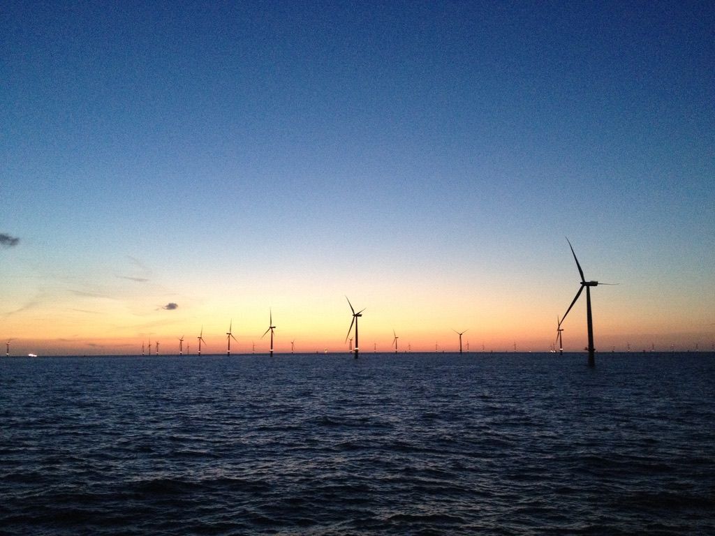 Offshore wind farm at sunset with wind turbines silhouetted against a colourful sky, overlooking calm ocean waters.