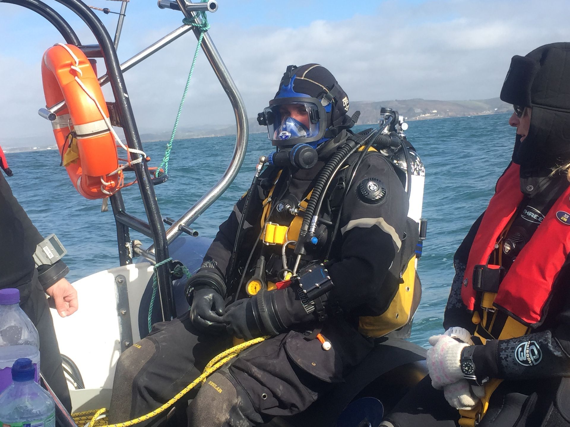 Two divers in full gear on a boat, one with a full-face mask preparing for a dive, while the other watches.