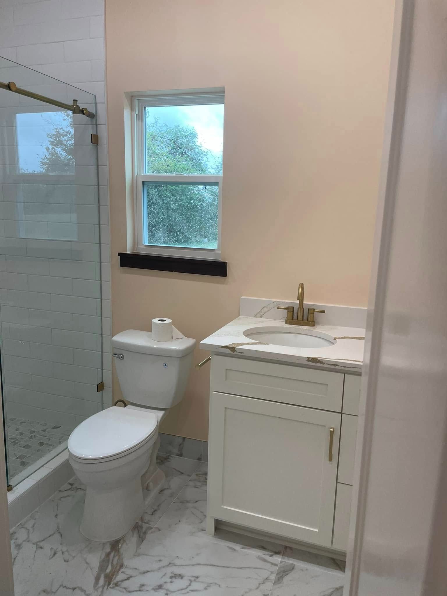 A small bathroom with a white toilet, vanity, and marble-look floor. A glass shower and a window with a dark sill are also visible.
