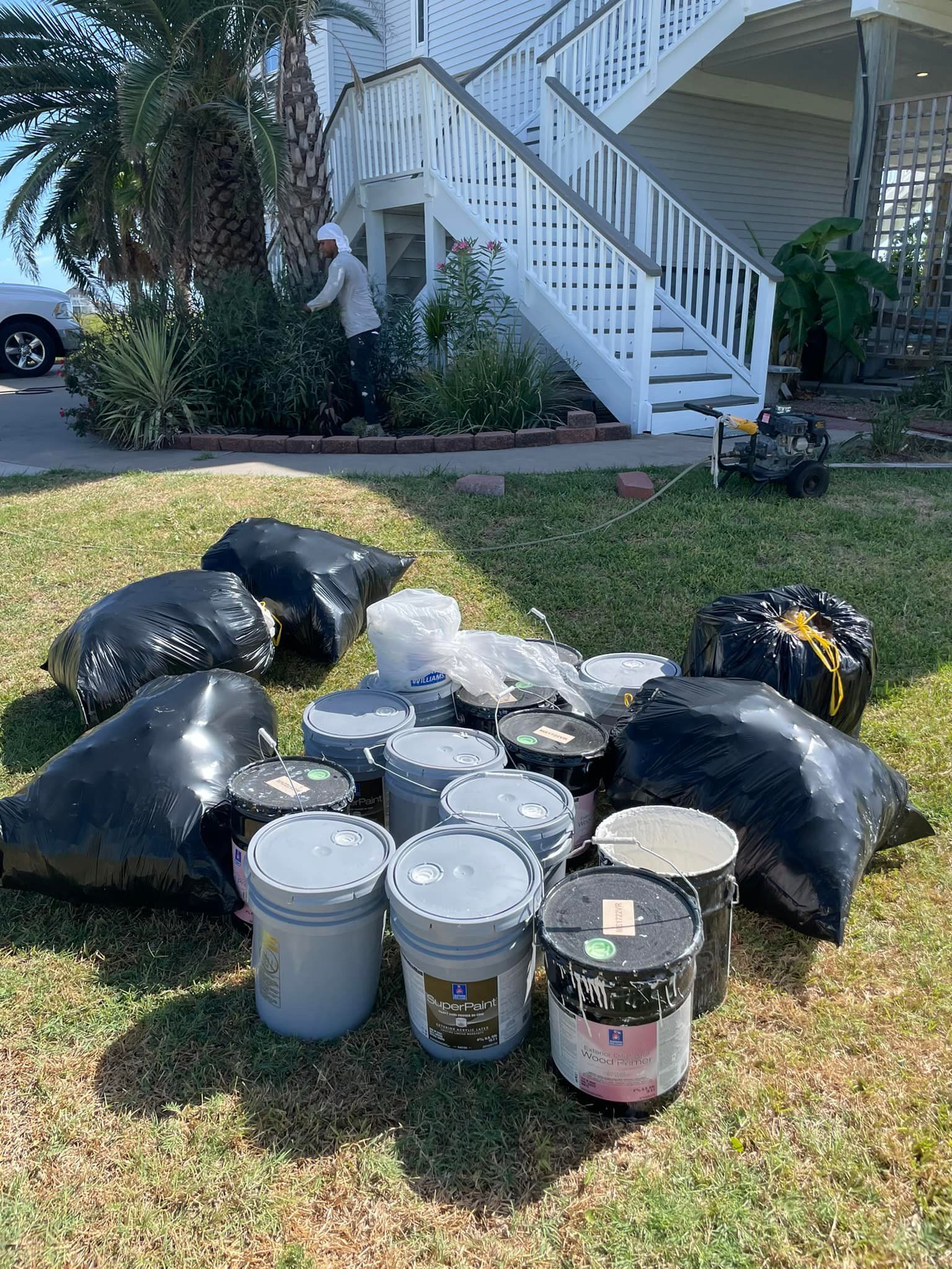 Person beside a white house with outdoor staircase and several black trash bags and buckets of paint on a green lawn.