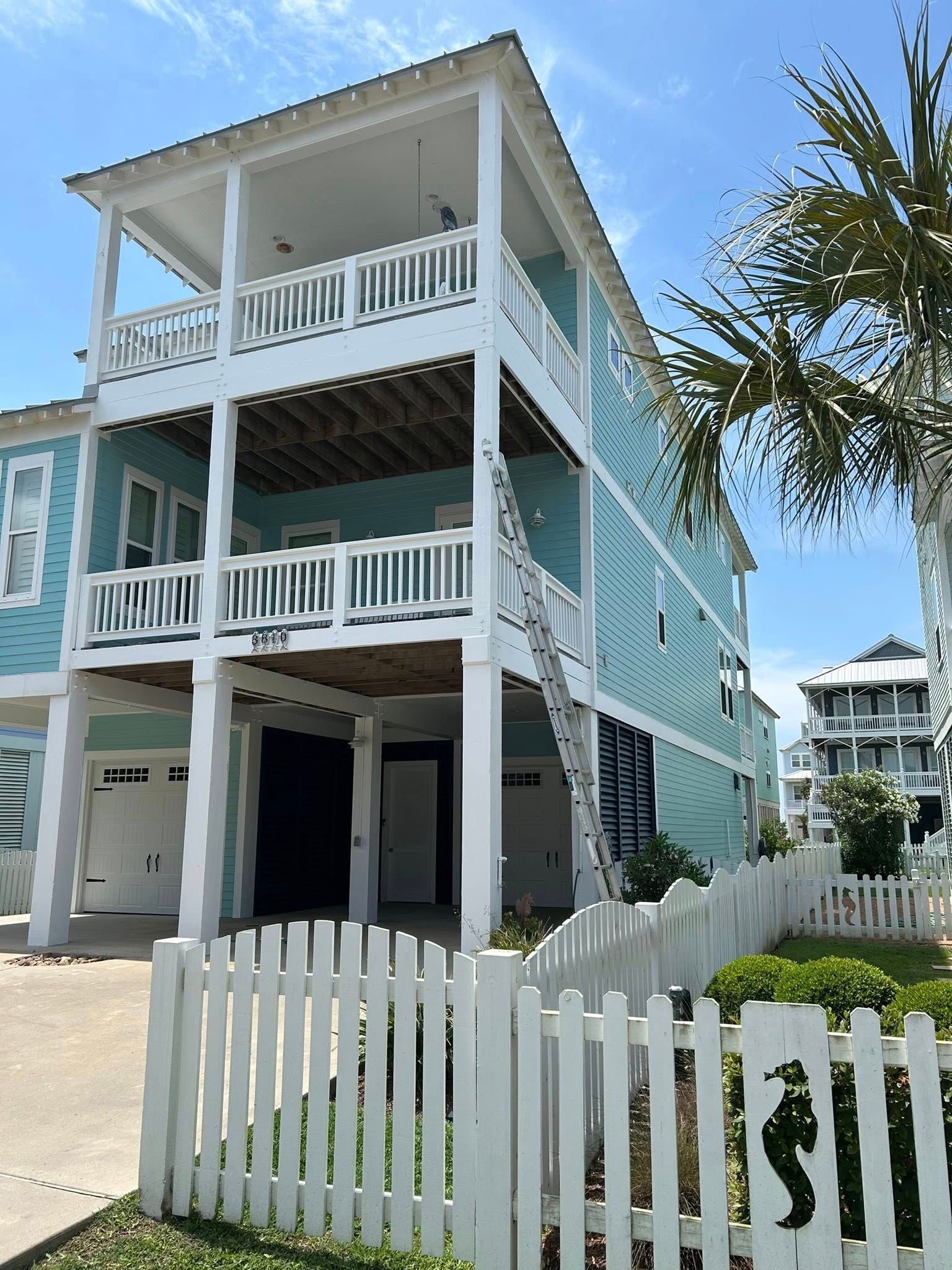 Three-story teal beach house with white railings and a white picket fence. A ladder leans against the side.
