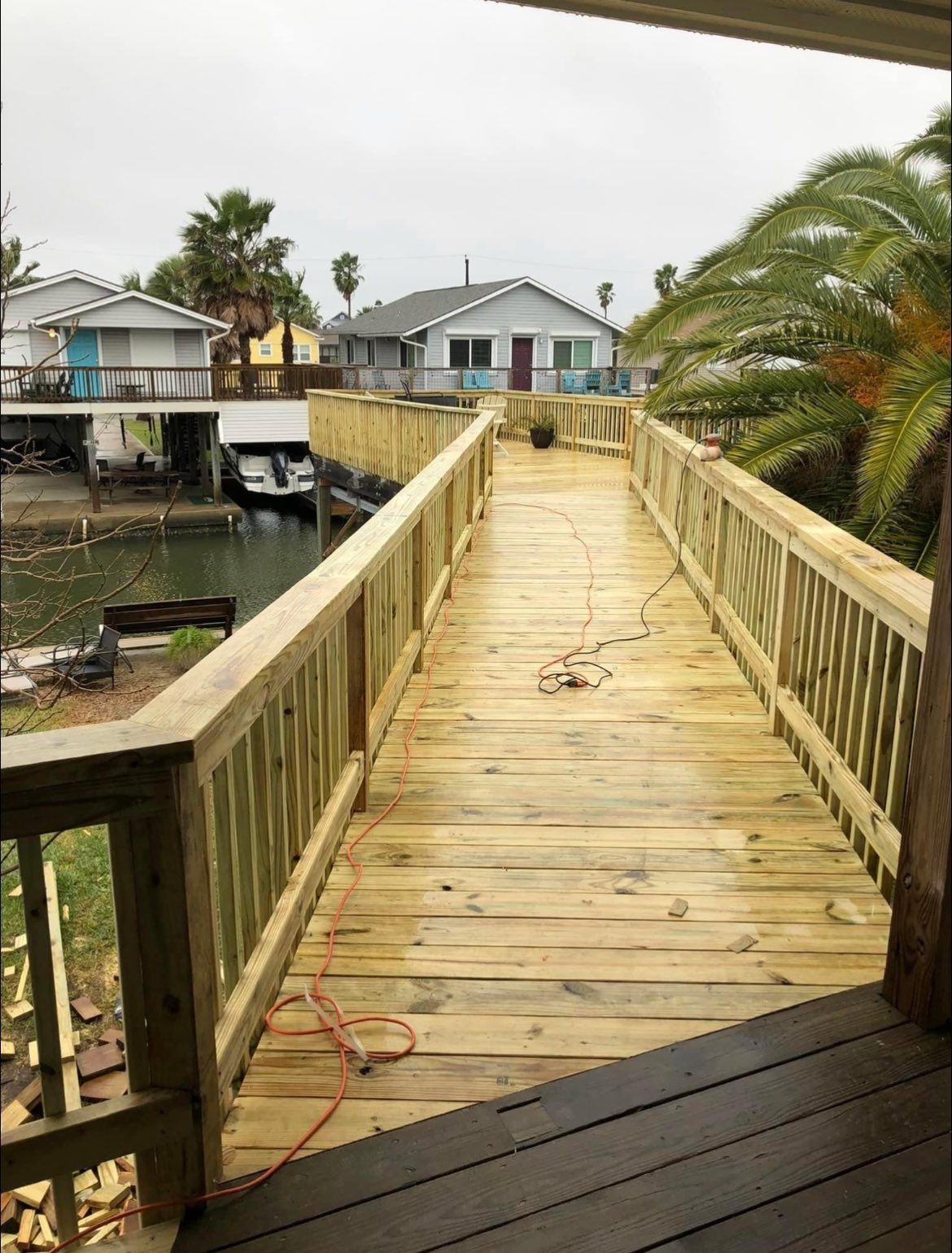 Wooden boardwalk extending over water, leading towards waterfront houses. Construction tools and materials visible.