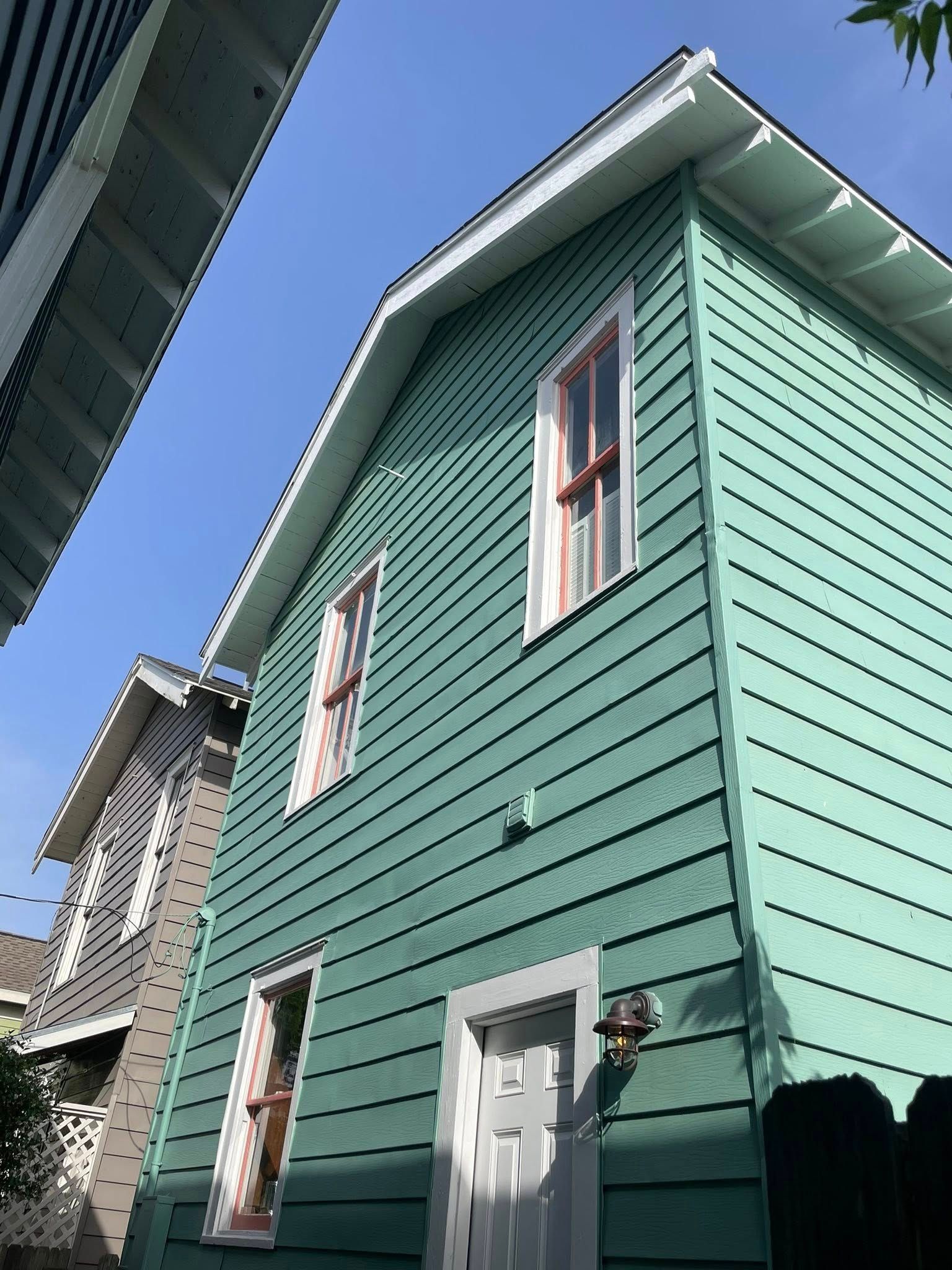 A two-story house with green siding, white trim, and windows with red frames, set against a blue sky.