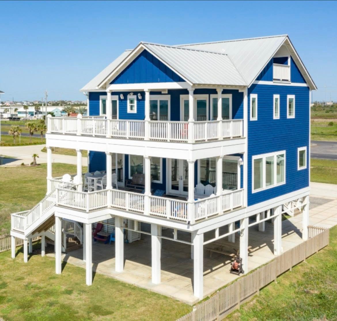 Two-story blue beach house with white trim, elevated on stilts. It has balconies, a white roof, and is set on a grassy lot.