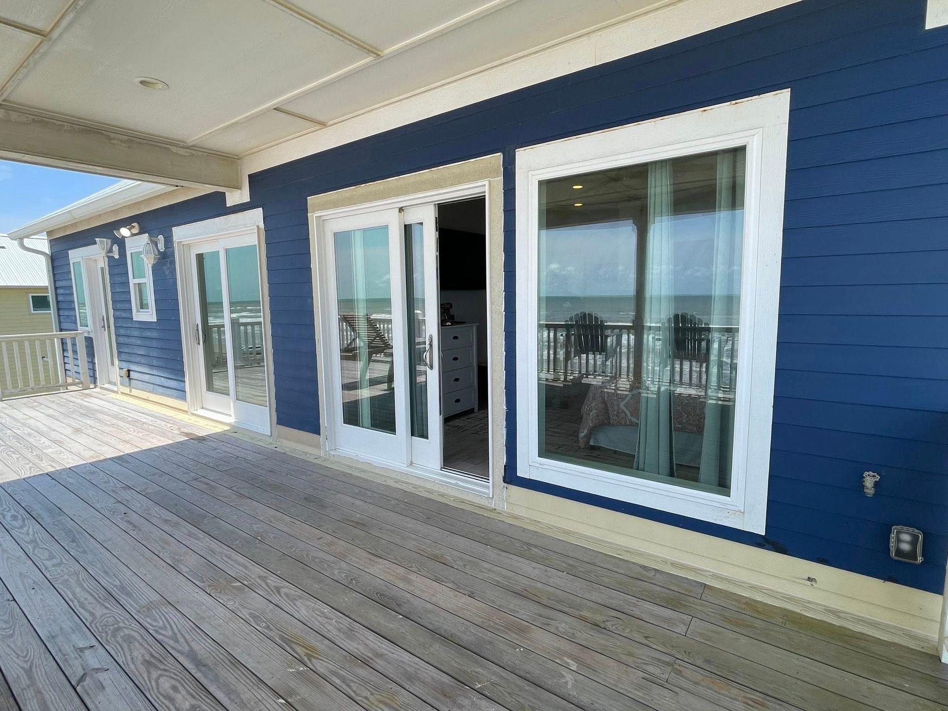 A blue beach house with white-framed windows and doors opens onto a wooden deck overlooking the ocean.