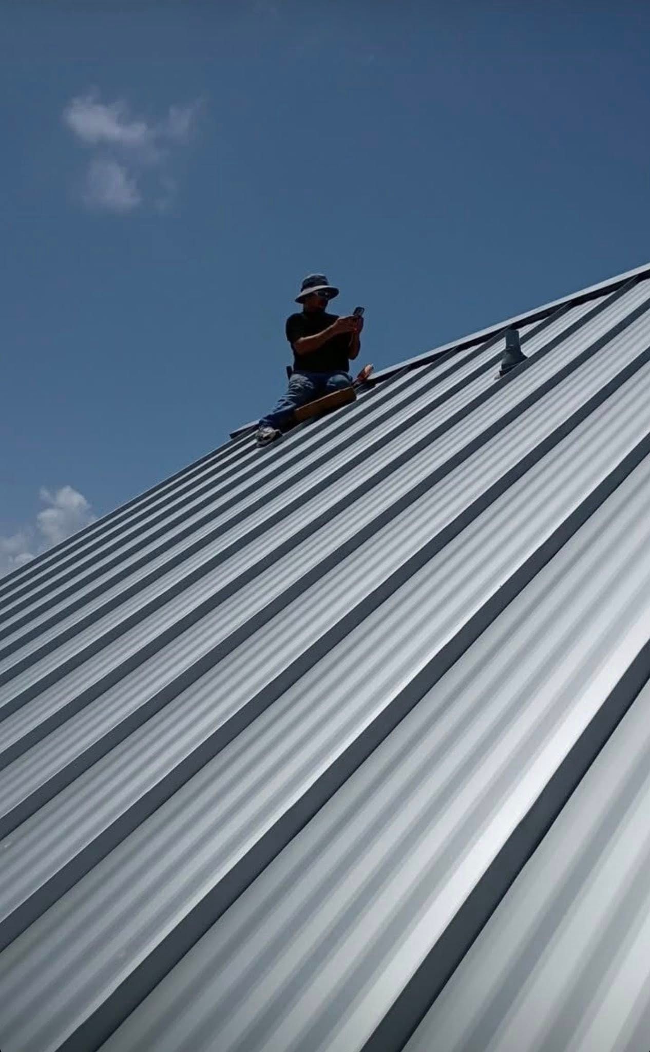 A person works on a metal roof under a blue sky. The roof has parallel ridges.