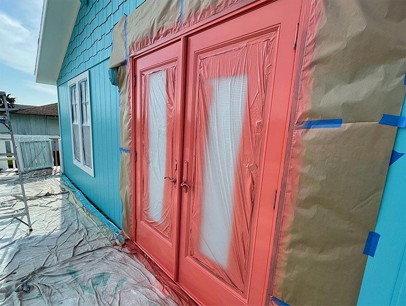 Person beside a white house with outdoor staircase and several black trash bags and buckets of paint on a green lawn.