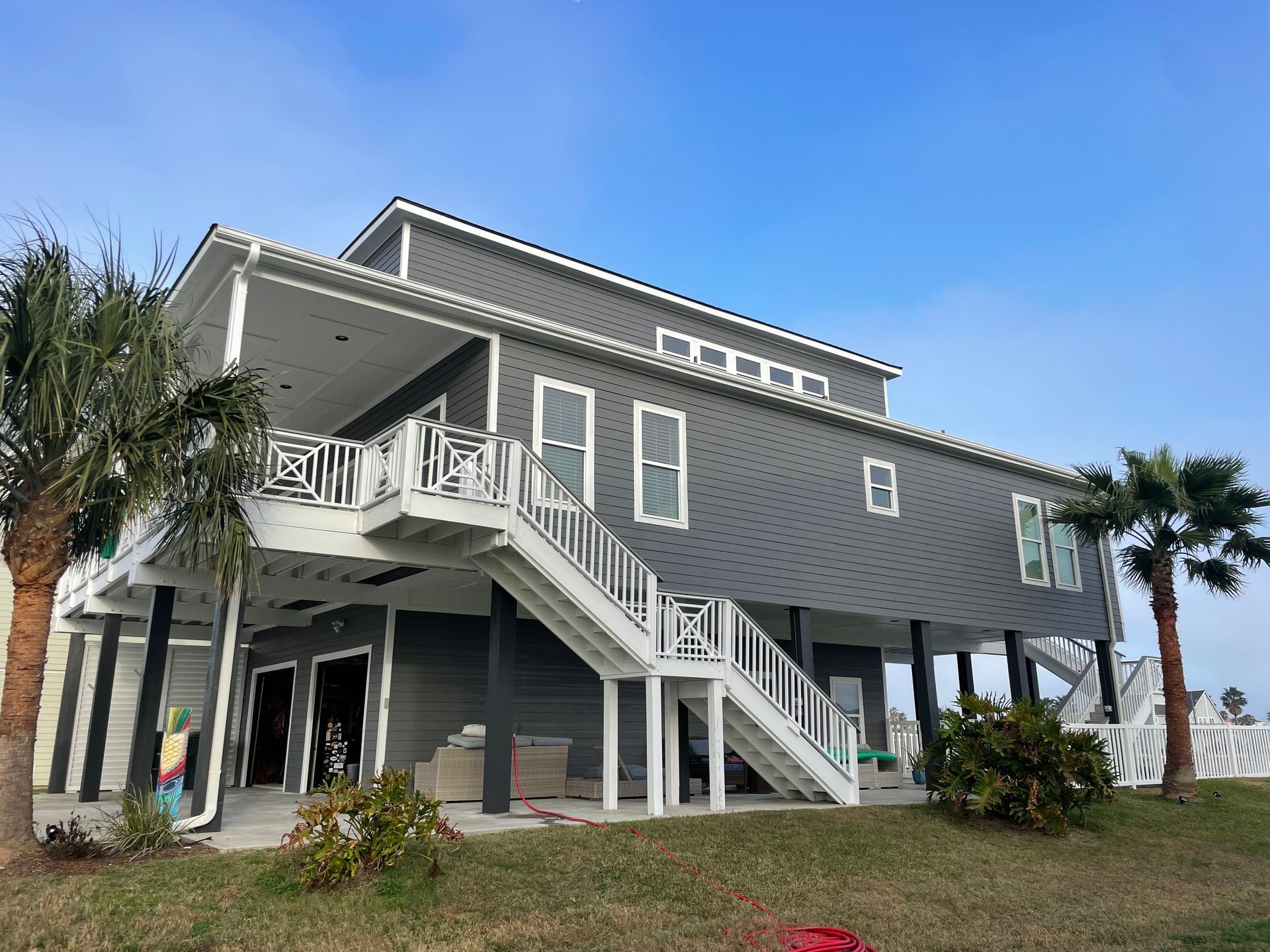 Two-story gray house on stilts with white trim, palm trees, and a blue sky. The house has a staircase and balcony.