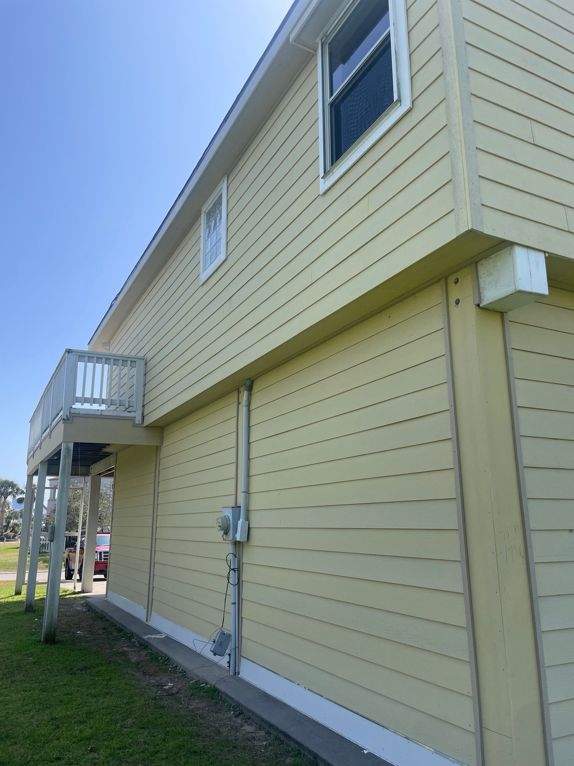 Side view of a two-story yellow house with a small balcony. The house has a concrete foundation and a blue sky in the background.